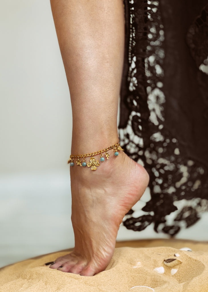 Gold chain anklet, with charms: one big gold flower and four small flowers and four turquoise round stones. Worn around a woman's ankle.
