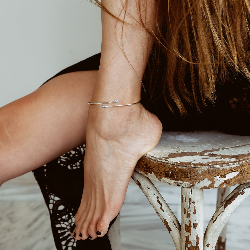 Γυναικείο πόδι που φοράει στον αστράγαλο ένα λεπτό χειροποίητο βραχιόλι ποδιού ασημί, με γαλάζιες πέτρες στις άκρες.