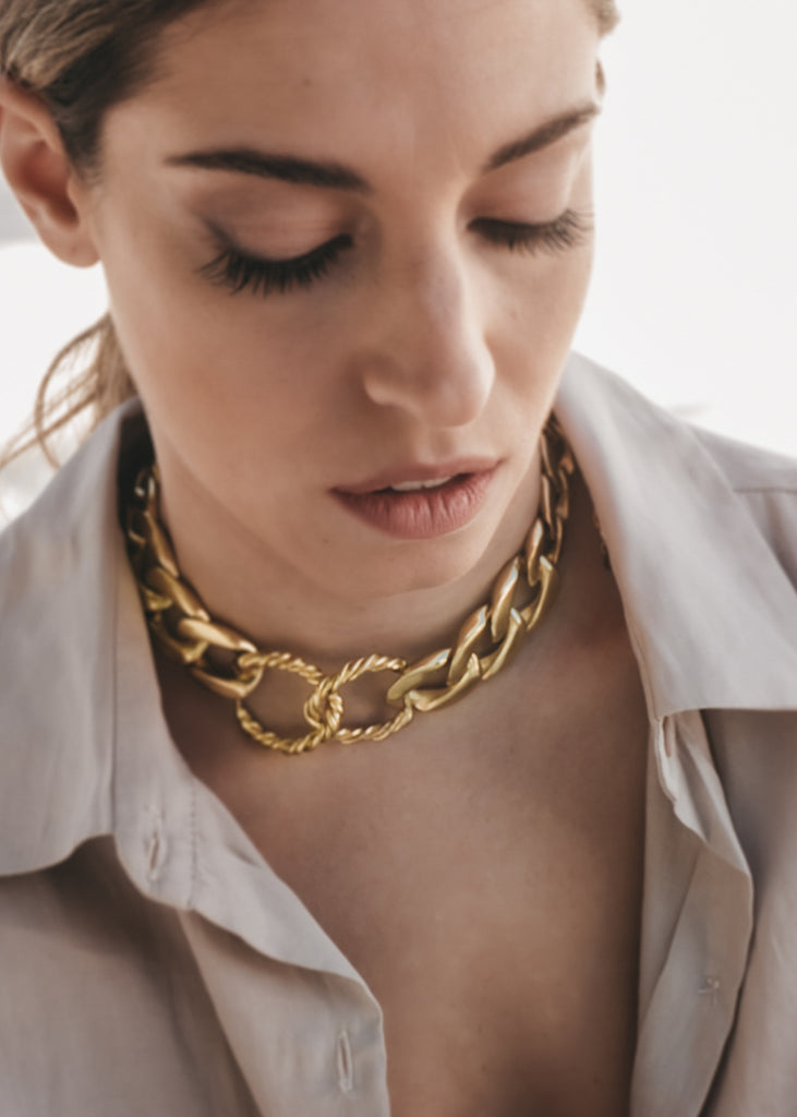Brunette looking down. She is wearing a beige, unbottuned shirt and a big, gold, link chain choker necklace