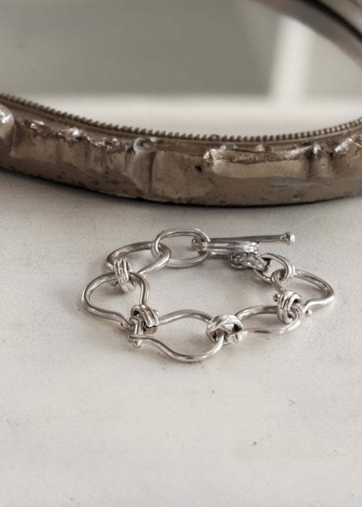 Silver, unique, link chain bracelet, placed on a white surface next to a vintage, brown mirror