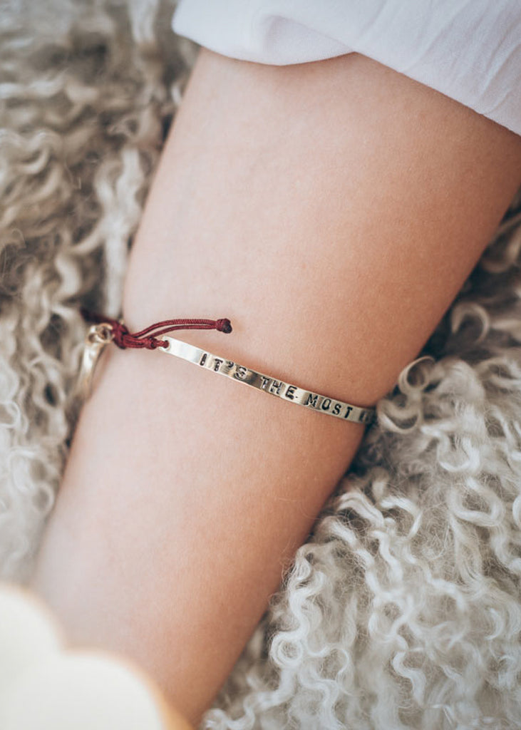Cropped photo of a forearm, on a fluffy pillow/carpet, wearing a red cord, adjustable, silver, charm bracelet