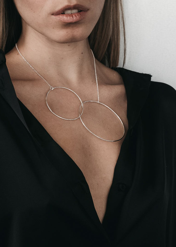 Lips to lower bust, cropped photo of female wearing a black, unbuttoned shirt. On her neck, a silver chain necklace with two interlocked link pendants