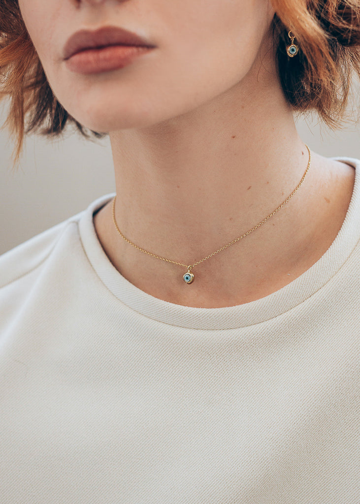 Close up of female's neck. She is wearing a white t-shirt and a gold, thin chain, evil eye necklace