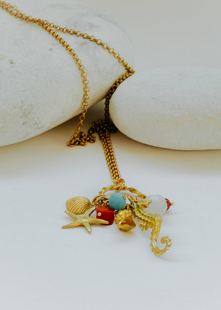 Long chain necklace in gold, with charms;  a starfish, two seashells, a seahorse and stones in red, turquoise and white. 