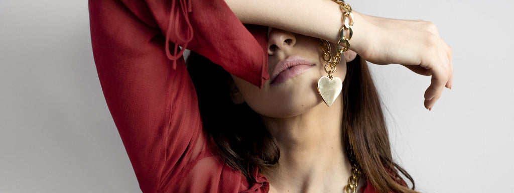 Female crossing her right arm over her eyes. On her wrist she is wearing a gold, chunkky chain bracelet with a large, heart pendant