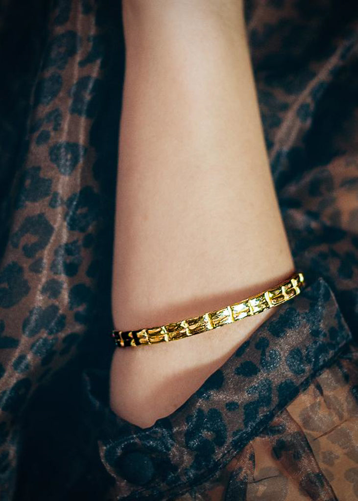 Close up of a left forearm. Semi transparent, patterned blouse, is rolled up. A gold bangle bracelet is worn