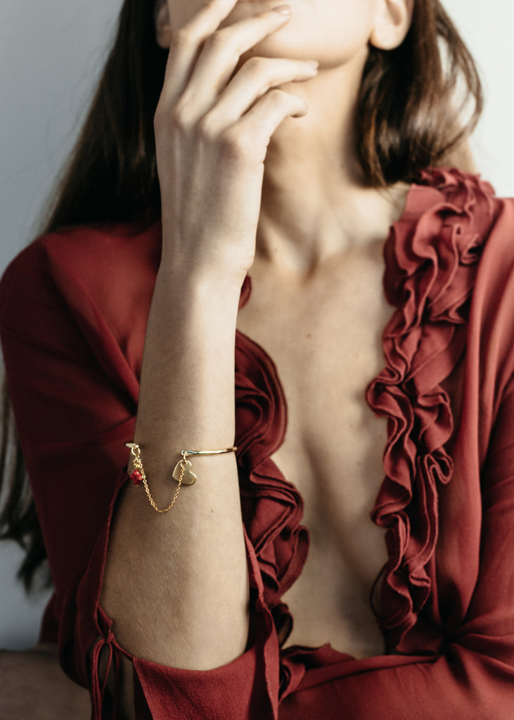 Female in a red, v-neck, unbuttoned, red, ruffled shirt. On her forearm, she is wearing a gold bangle, and chain bracelet by 3rd Floor