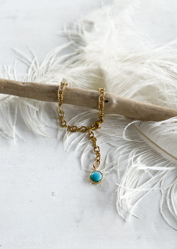 Babbett. Gold plated anklet, with a turquoise stone, photographed laying on a white feather