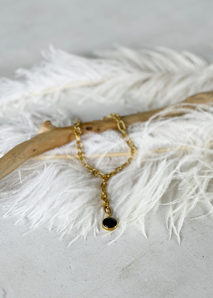 Babbett. Gold plated anklet, with a black onyx stone, photographed laying on a white feather.Χειροποίητο Βραχιόλι Ποδιού Babbett,  με μαύρη πέτρα