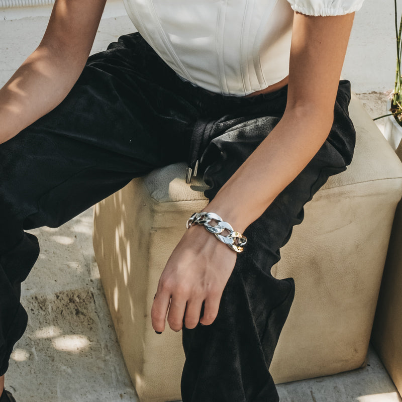 Female in black trousers sitting on a beige stool. On her wrist she is wearing a silver, chunky chain bracelet
