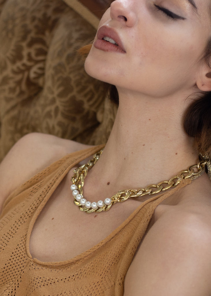 Young female resting back on a couch. Her eyes closed. She is wearing a gold, link chain, and pearls necklace and a brown summer blouse