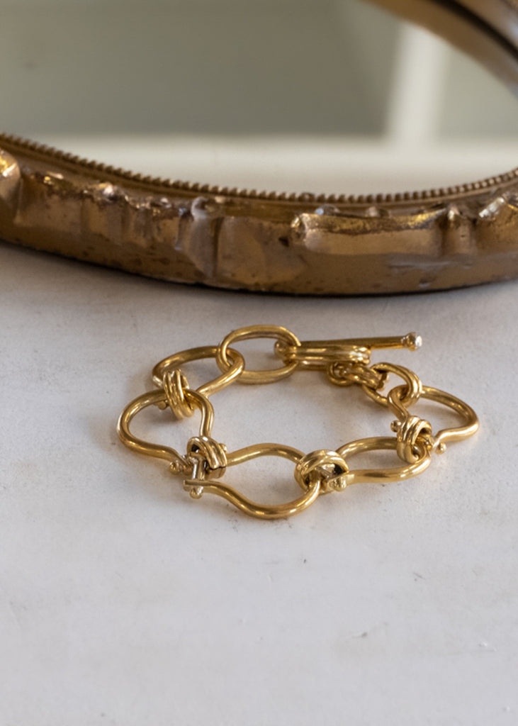 Gold, unique, link bracelet, placed on a white surface, next to a mirror. 