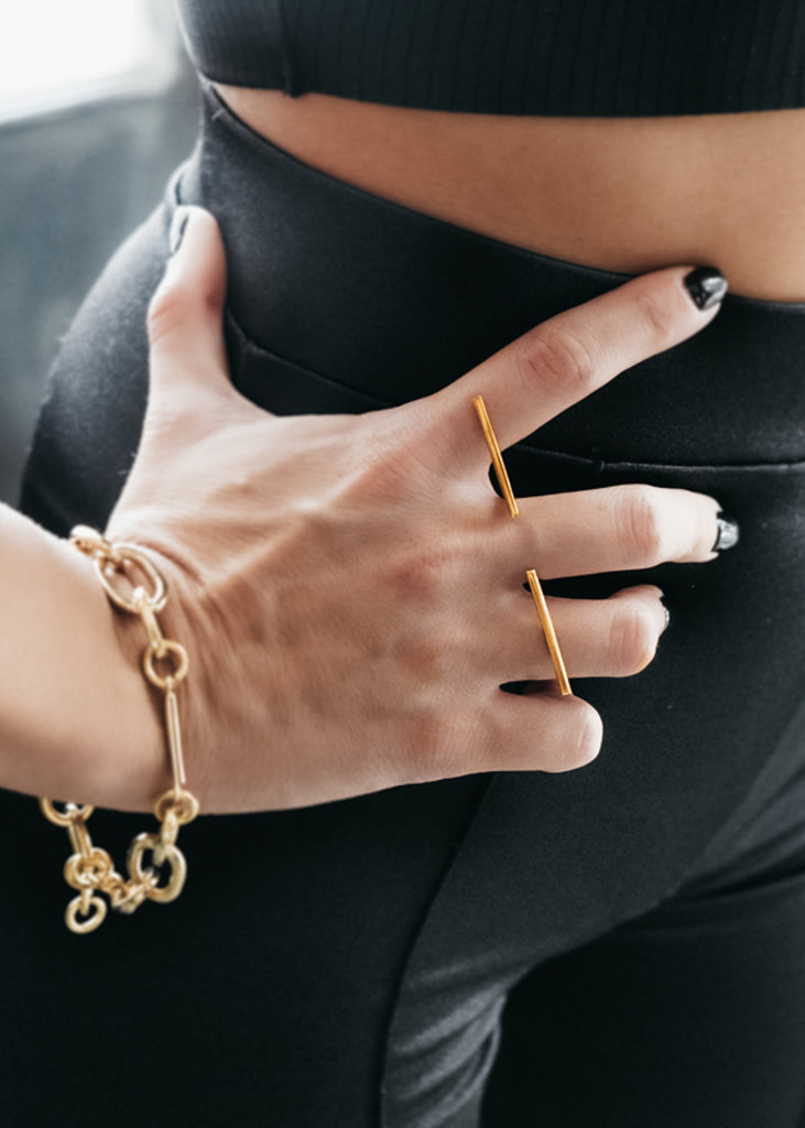 Female in total black, with her right hand on her hip. She is wearing a gold, adjustable, stick ring and a gold bracelet