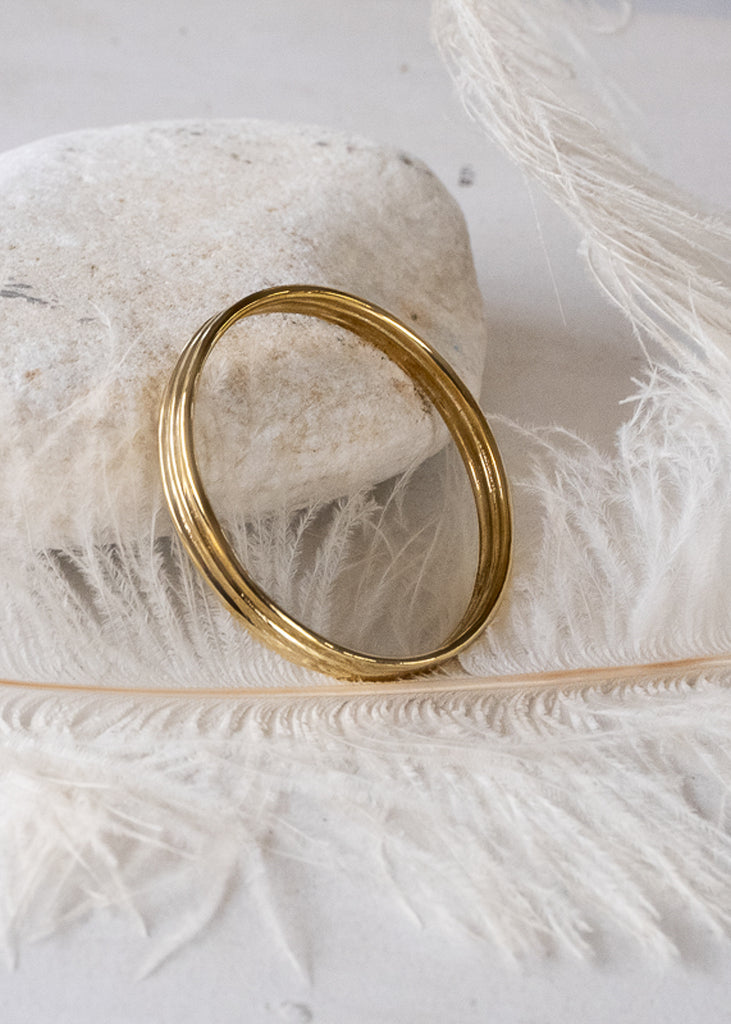 Vesper. Gold, bangle bracelet placed on a white rock, and on a white feather