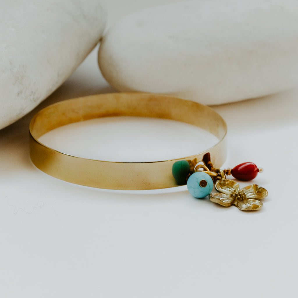 Gold wide bracelet with three charms, a gold flower, a round turquoise stone and a red bead.