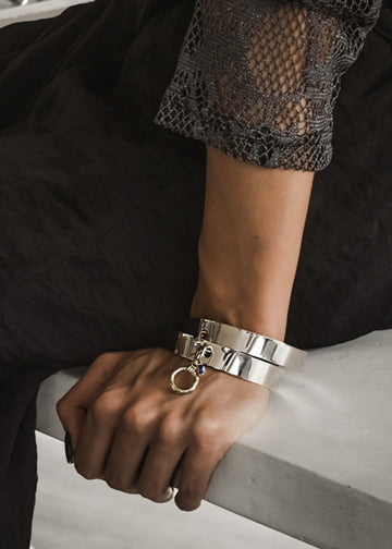 Woman in dress black. She is sitting on a white bench. On her left wrist, she is wearing a Vogue, double bangle, silver plated brass, handmade bracelet