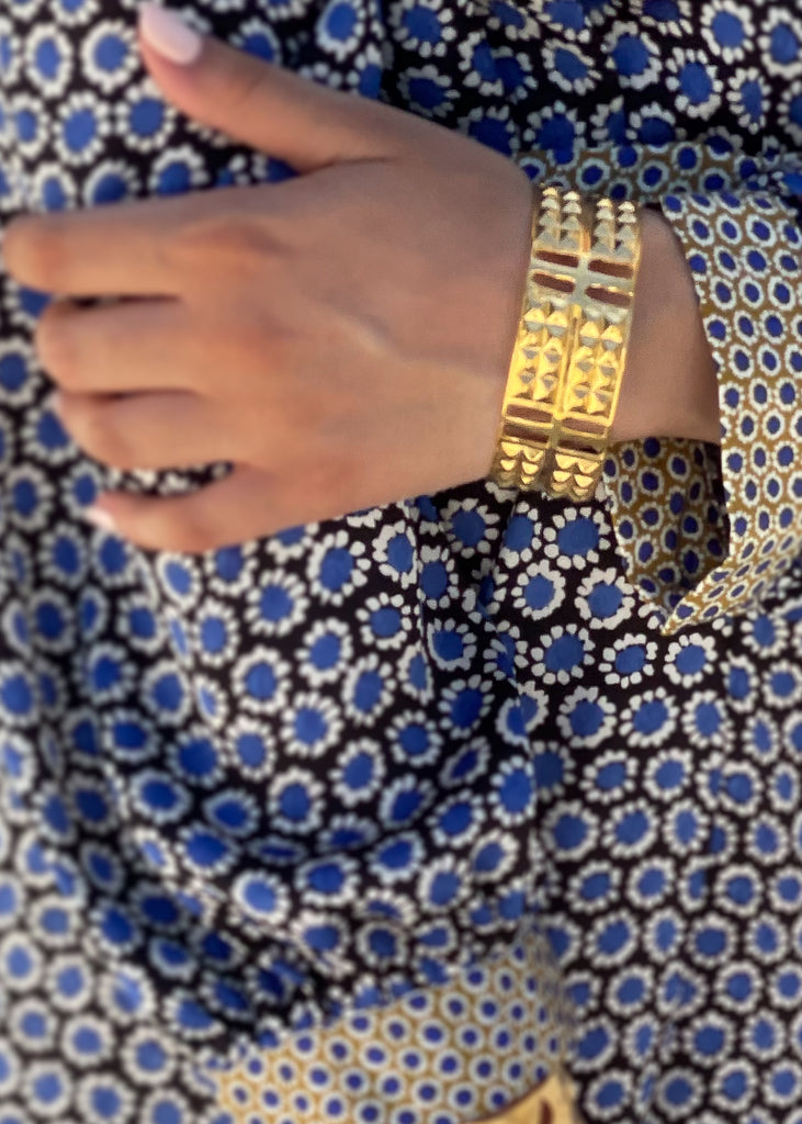 Female's arm, in a blue dress, leaned over the back of a chair. She is wearing a gold, statement cuff bracelet