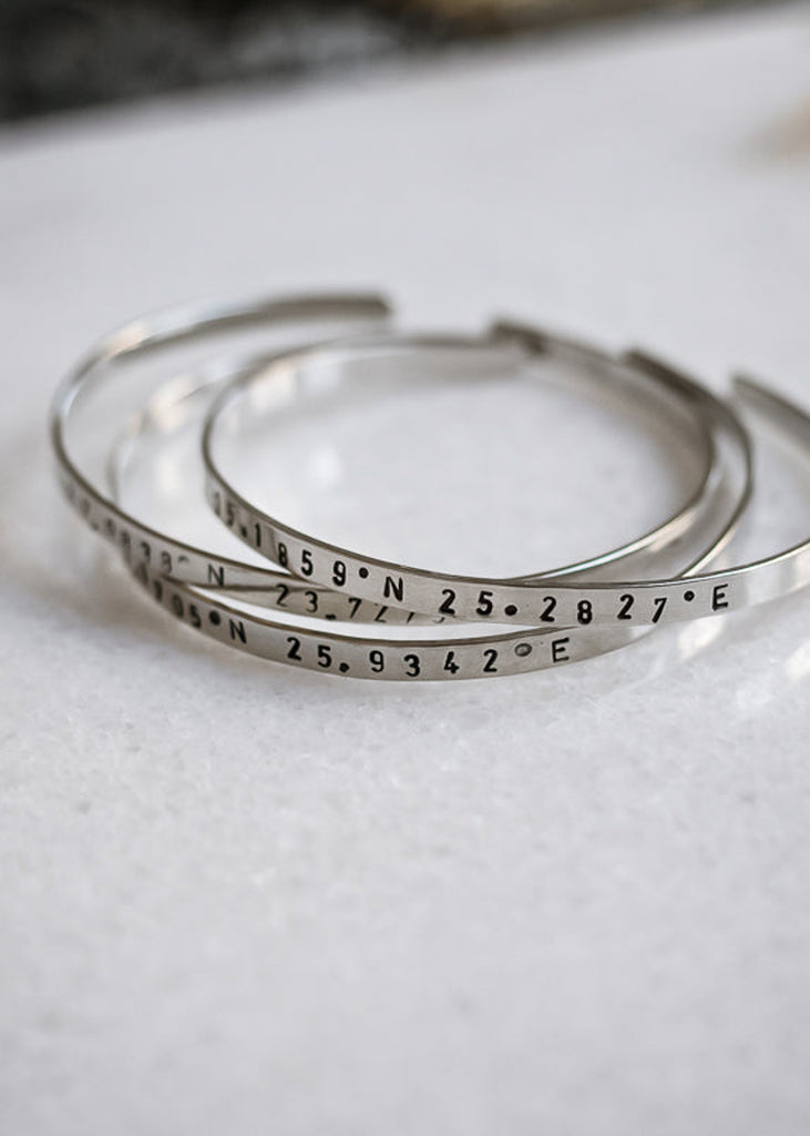 Three, adjustable, silver bracelets  stamped with geographical coordinates, placed on a white marble surface.