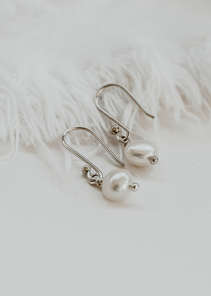 Two silver earrings with one small pearl hangind on each placed on a white background.