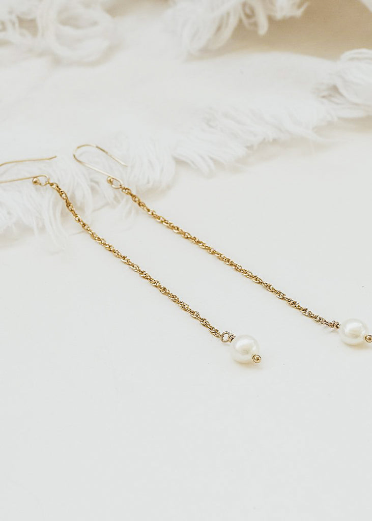 Two gold dangling earrings, with a thin long chain and a round pearl on each end, placed on a white background. 