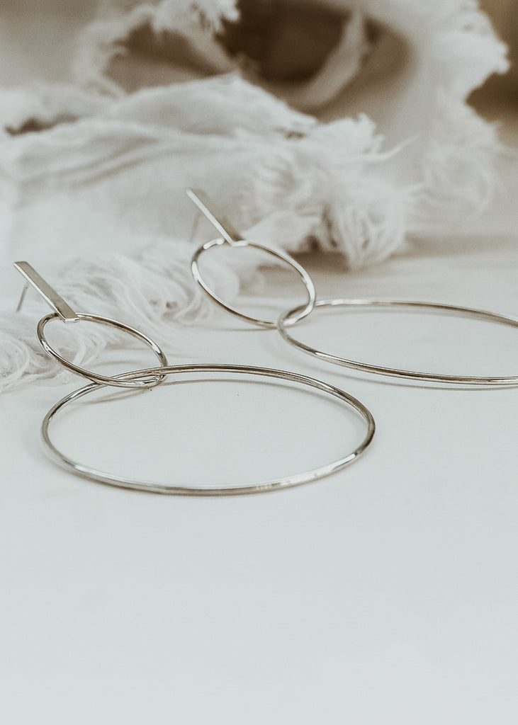 Silver dangling earrings made of two uneven thin circles of sterling silver placed on a table. | Χειροποίητα Σκουλαρίκια Mona Ασημί
