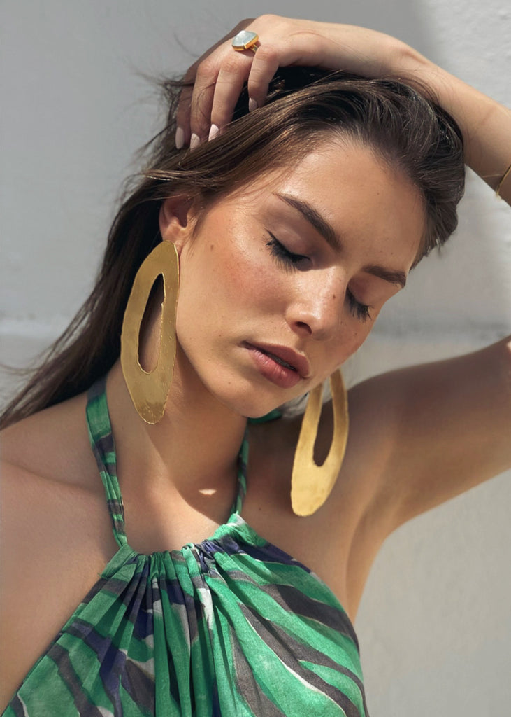 woman in a summer dress with her eyes closed and her arm holding her hair is wearing the statement earrings Toma in gold 