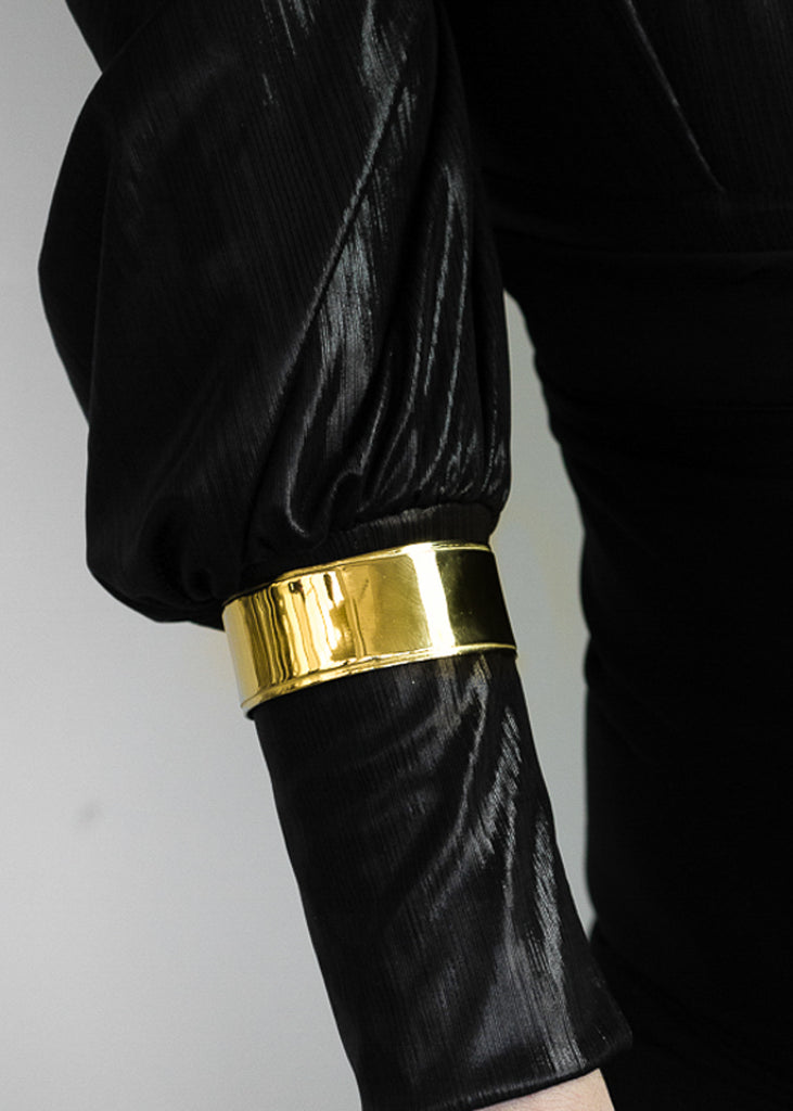 Photo of what appears to be a female's arm, in a black cuff sleeve shirt. Over the sleeve she is wearing a gold cuff bracelet