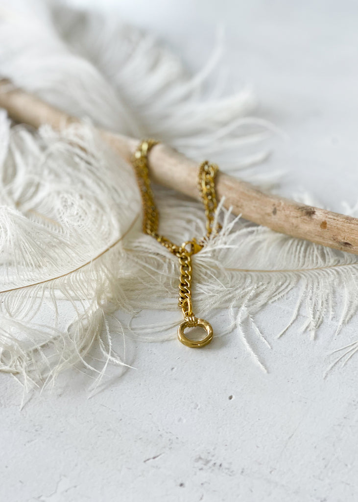 Gold anklet, with a dangling, gold link, artistically photographed on a piece of wood and white feathers