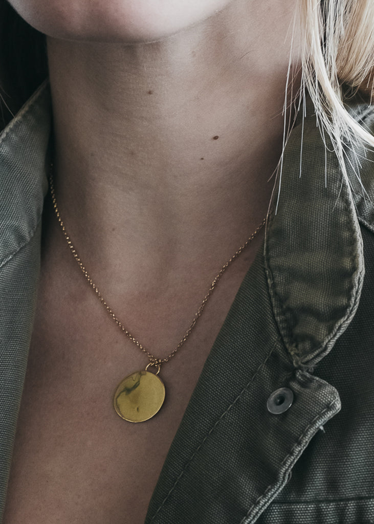 Extreme close up of a female's chest. She is wearing a gold, thin chain necklace, with a flat, round, gold pendant. By 3rd Floor Handmade Jewellery