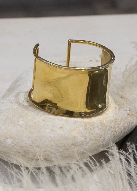 Fallon. Gold plated, wide, statement cuff, placed on a white rock, which is in turn placed on a white feather