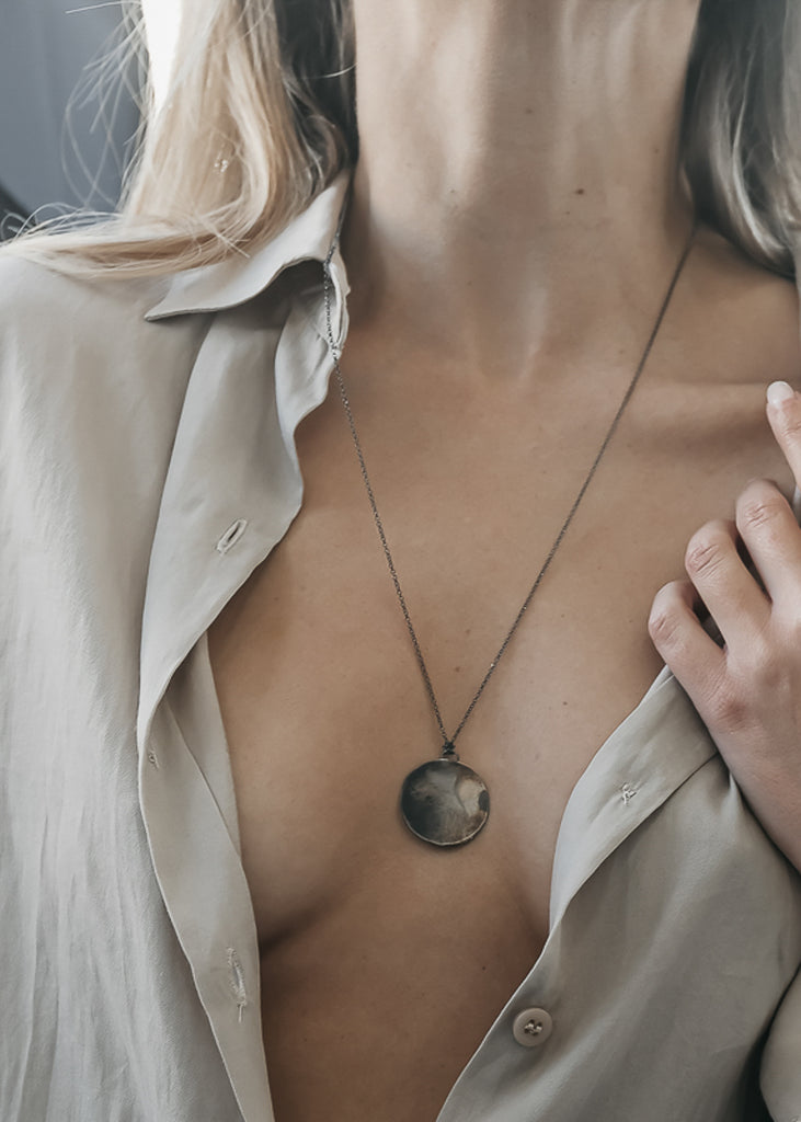 Female in unbuttoned grey shirt. On her neck, a long, thin chain necklace with a flat, round pendant