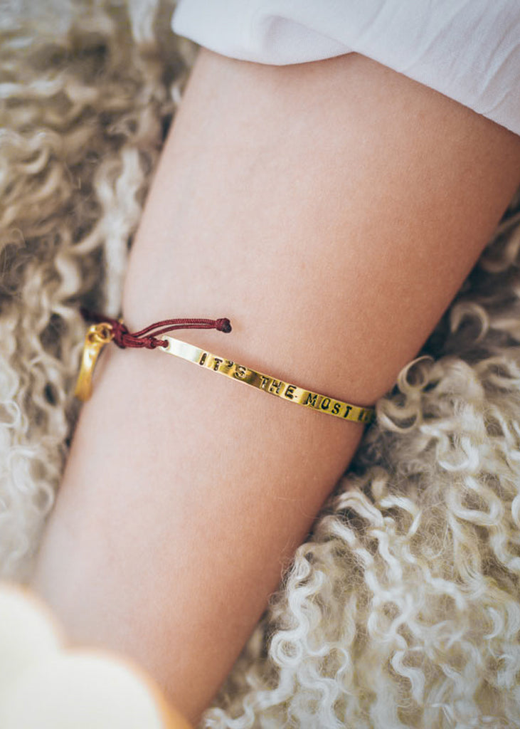 Cropped photo of a forearm, on a fluffy pillow/carpet, wearing a red cord, adjustable, gold, charm bracelet