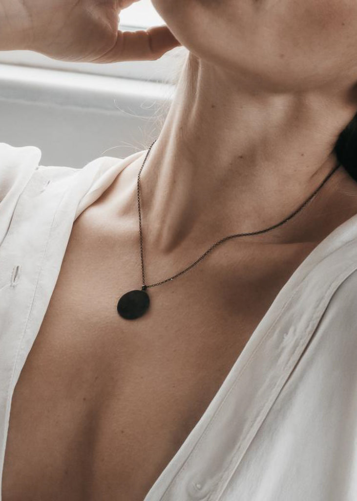 Close up of female's neck. She is wearing a black chain necklace with a thin, round, black, pendant.