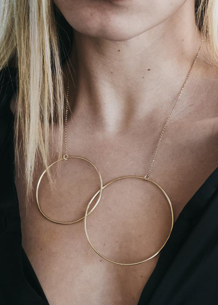 Chin to bust, cropped photo of female in a black, unbuttoned shirt. On her neck she is wearing a gold chain necklace, with two, interlocked link pendants.