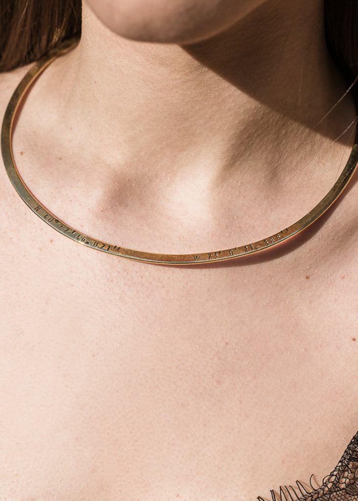 Girl in black top, wearing a flat gold plated collar neckpiece stamped with longitudtude coordinates-gold