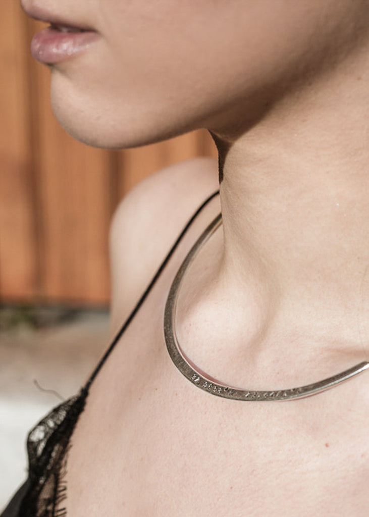 Girl in black top, wearing a flat gold plated collar neckpiece stamped with longitudtude coordinates