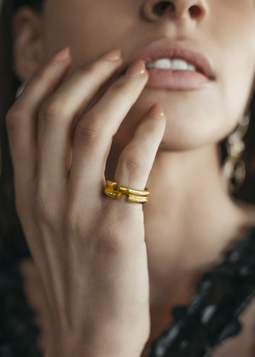 Close up of a female's hand. Her fingers gently touching the edge of her mouth. On her pinkie, she is wearing a double rod, gold ring