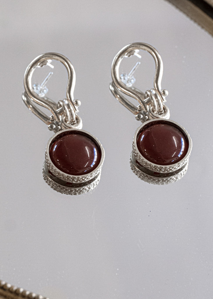 Temptress. Unique link, pendant earrings with a round, brown carnelian stone. Earrings are placed on a mirror